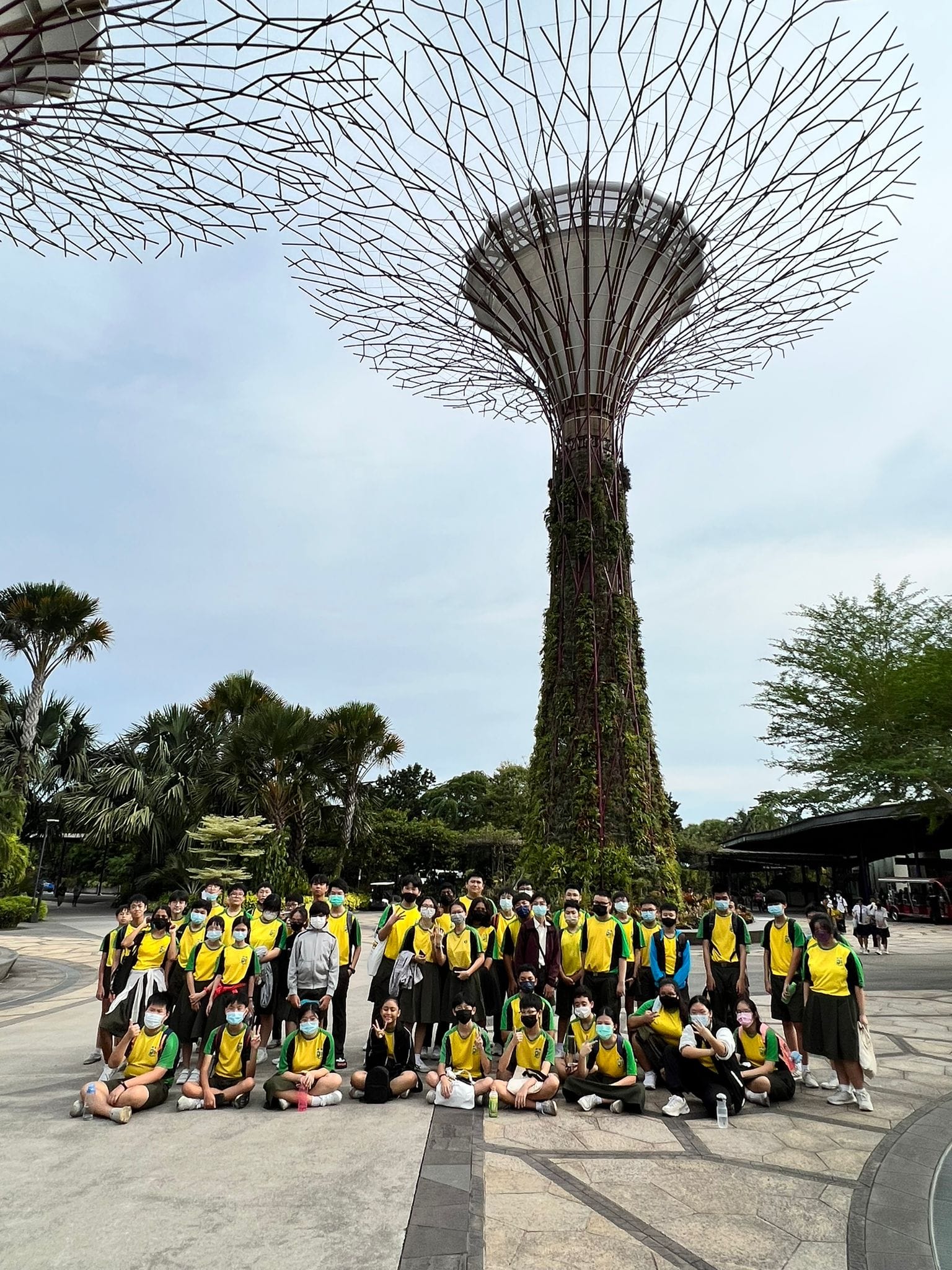 Group in yellow shirts posing in front of a tall metal tree tower with branching frame and greenery.